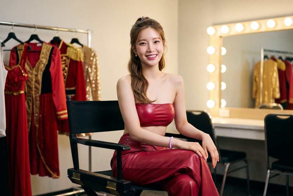 Sela Moon smiling backstage on a director’s chair beside a rail of red and gold stage outfits